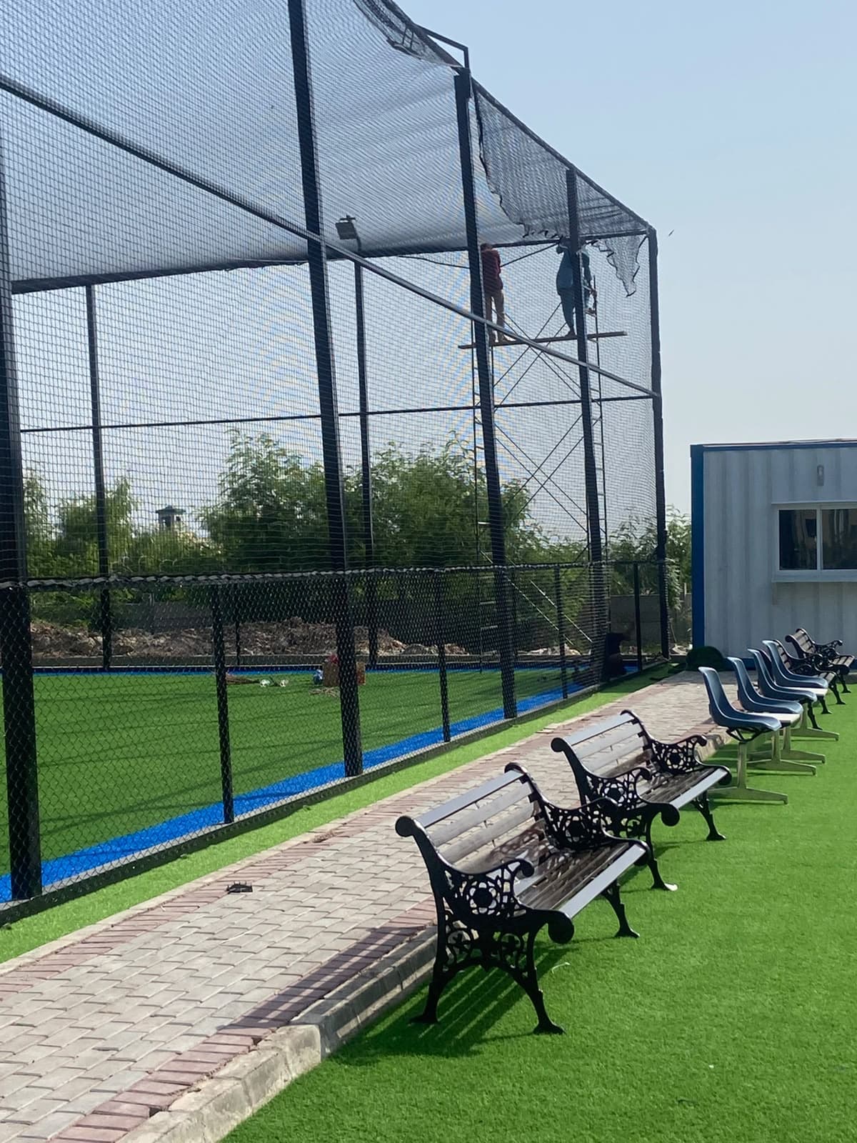 Spectator concrete bench seating, interlocking paver walkway, and scaffolding crew securing perimeter netting at Football Ground Askari 14